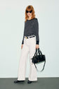 Woman with red hair, wearing black sunglasses, a T-shirt à rayures coupe ajustée à manches longues, white wide-leg pants, and black shoes, stands holding a black handbag against a plain light background.