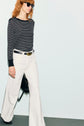 Person wearing a T-shirt à rayures noir et blanc à manches longues, white wide-leg pants, a black belt, sunglasses, and holding a black bag, walking against a light background.