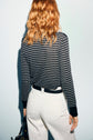 A person with wavy, shoulder-length red hair is shown from the back, wearing a T-shirt à rayures noire et blanche à manches longues and white pants in a coupe ajustée, holding a black bag by their side.