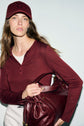 A woman wearing a t-shirt bordeaux and matching cap holds a large burgundy leather bag. She has long brown hair and is standing against a plain light background.