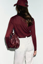 A woman with long brown hair wears a fine knit burgundy cap, a t-shirt bordeaux, and white pants, holding a matching burgundy handbag over her shoulder. She is facing away from the camera.