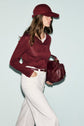 Woman in a t-shirt bordeaux and cap carries a matching handbag, wearing wide-leg white pants and a white belt, walking against a light blue background.