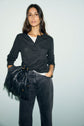 A woman with long brown hair wears a t-shirt anthracite and black jeans, holding a black fringed handbag. She stands against a light background, looking confidently at the camera.