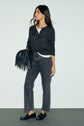 Woman in a dark long-sleeve maille fine top, gray jeans, and black loafers stands holding a black fringed handbag, looking to the side against a plain light background.