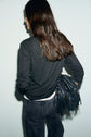 A person with long brown hair, wearing an anthracite t-shirt and jeans, stands facing away, holding a black fringed bag over one shoulder against a light background.