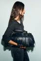 A woman with long brown hair wearing a t-shirt anthracite and a dark outfit holds a large black fringed handbag under her arm, standing against a plain light-colored background.