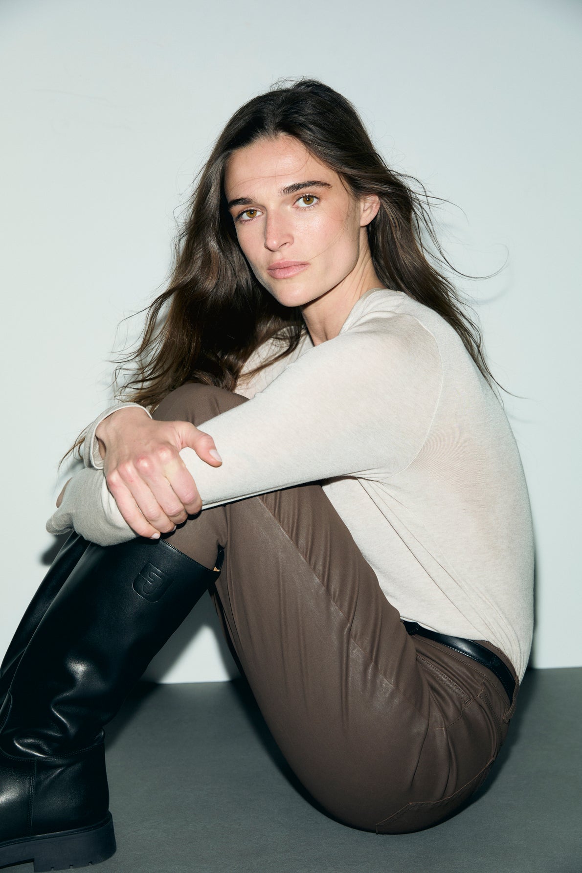 A woman with long brown hair sits on the floor against a white wall, wearing a tee-shirt beige en lyocell et laine with a col V satin, paired with brown pants and black boots. She looks at the camera with a neutral expression.
