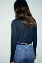 A person with long brown hair, wearing a black long-sleeve tee-shirt lyocell laine and high-waisted blue jeans with a black belt, stands facing away from the camera against a plain light background.