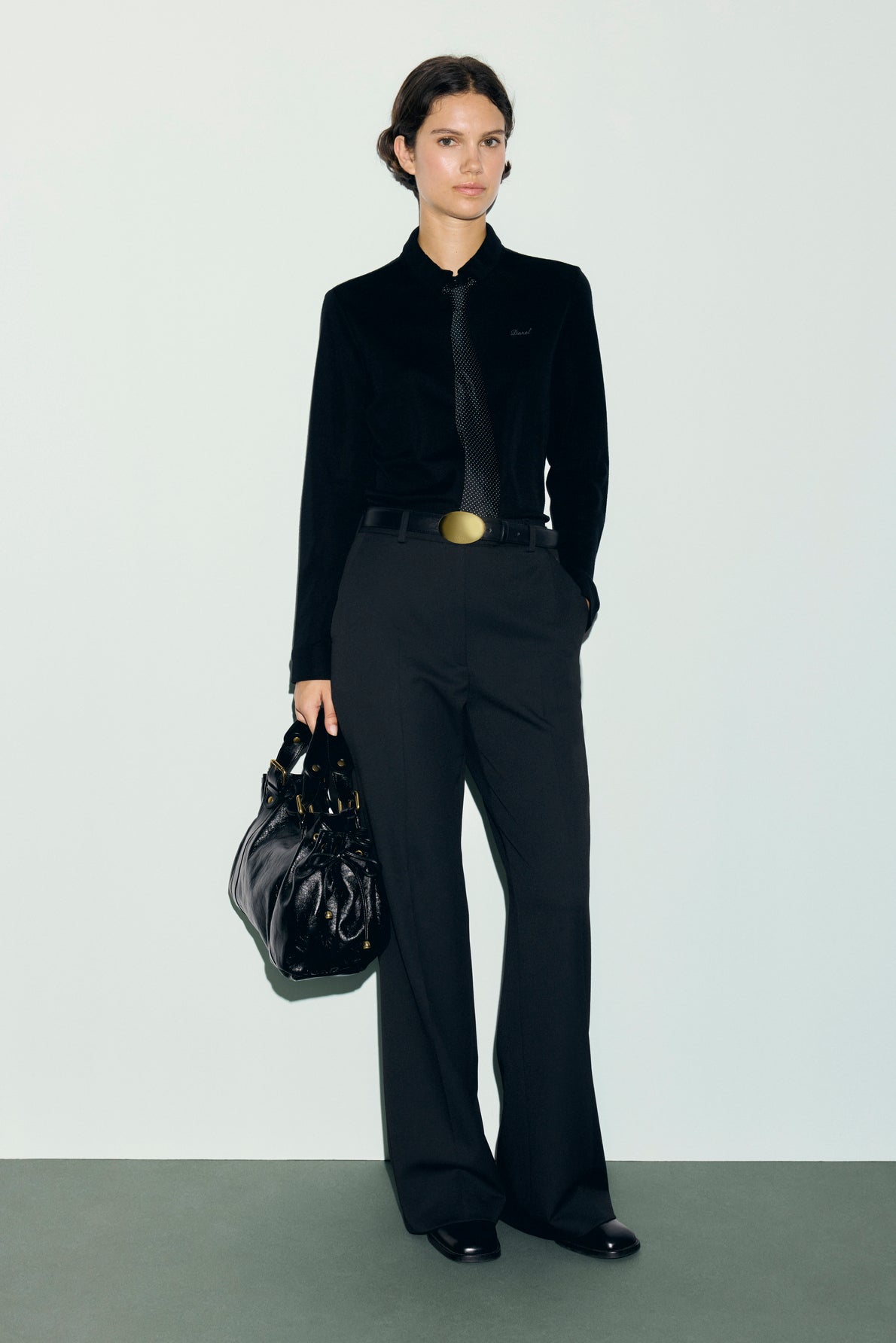 A person stands against a plain background wearing a chemise noire femme—a chic, black button-up shirt—styled with a matching black tie, high-waisted black trousers, and sleek black shoes. They hold a shiny black bag, and their hair is pulled back neatly for an effortlessly polished look.