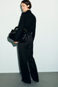 A person with short hair, dressed in a chemise noire femme and wide-leg pants, stands against a pale wall, looking over their shoulder and holding a large, shiny black handbag.