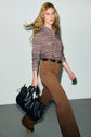 A woman with long blonde hair wears a chemise imprimée and brown pants, holding a large black handbag while walking confidently against a plain background.