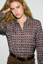 A woman with long wavy blonde hair poses in a chemise imprimée à manches longues, featuring a geometric pattern of blue and brown shapes. She wears brown pants with a navy blue belt, looking directly at the camera with one hand raised to her hair.