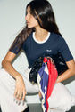 A woman with long brown hair wears a tee-shirt bleu marine en coton with a col contrasté and white pants, holding a black bag decorated with a colorful scarf while looking to the side.
