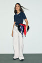 Woman with long hair wears a tee-shirt bleu marine en coton with a col contrasté, paired with white wide-leg pants and black shoes. She holds a black bag and a colorful scarf, standing against a plain light background and looking confidently at the camera.