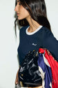 Woman with long dark hair in a navy tee-shirt bleu marine en coton with a subtle col contrasté holds a shiny black bag adorned with colorful scarves. She is shown in profile against a plain white background.