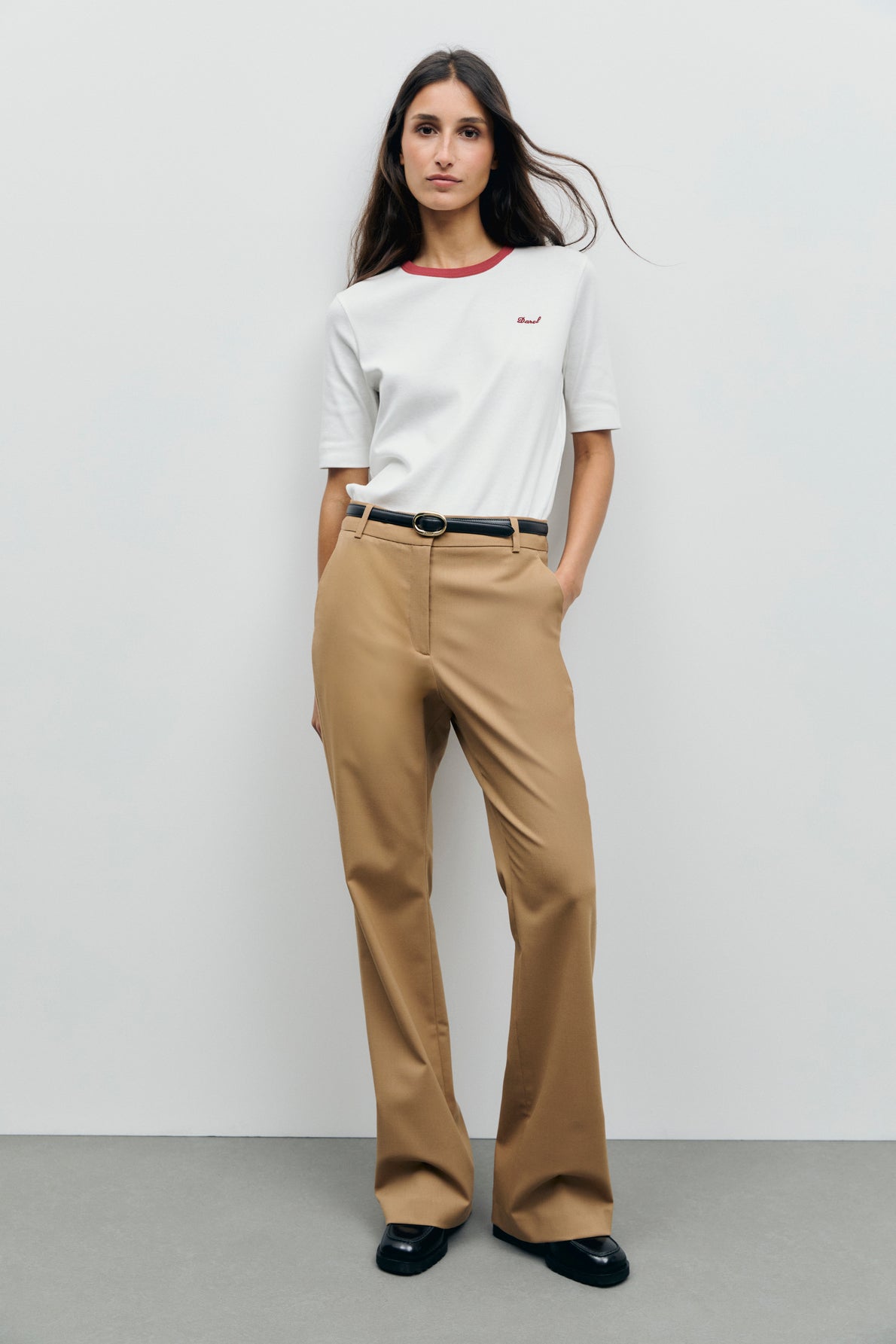 A woman stands against a plain background wearing a white short-sleeve tee-shirt en coton with red col contrasté, tan high-waisted trousers, a black belt, and black shoes. She has long brown hair and hands in her pockets.
