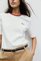 A person wearing a tee-shirt coton with col contrasté and motif brodé, tucked into tan pants with a black belt, stands against a plain light background.