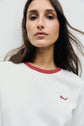 A woman with long brown hair wears a white tee-shirt coton with a col contrasté in red. The word Darcel is motif brodé in red on the shirt. She is looking directly at the camera against a plain background.