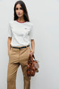 A woman stands against a plain light background, wearing a tee-shirt coton with motif brodé and red col contrasté, paired with tan trousers and a black belt, and holding a brown leather handbag.