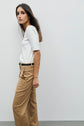 Woman with long brown hair wearing a white tee-shirt coton with col contrasté and tan high-waisted trousers, standing sideways against a plain white background.