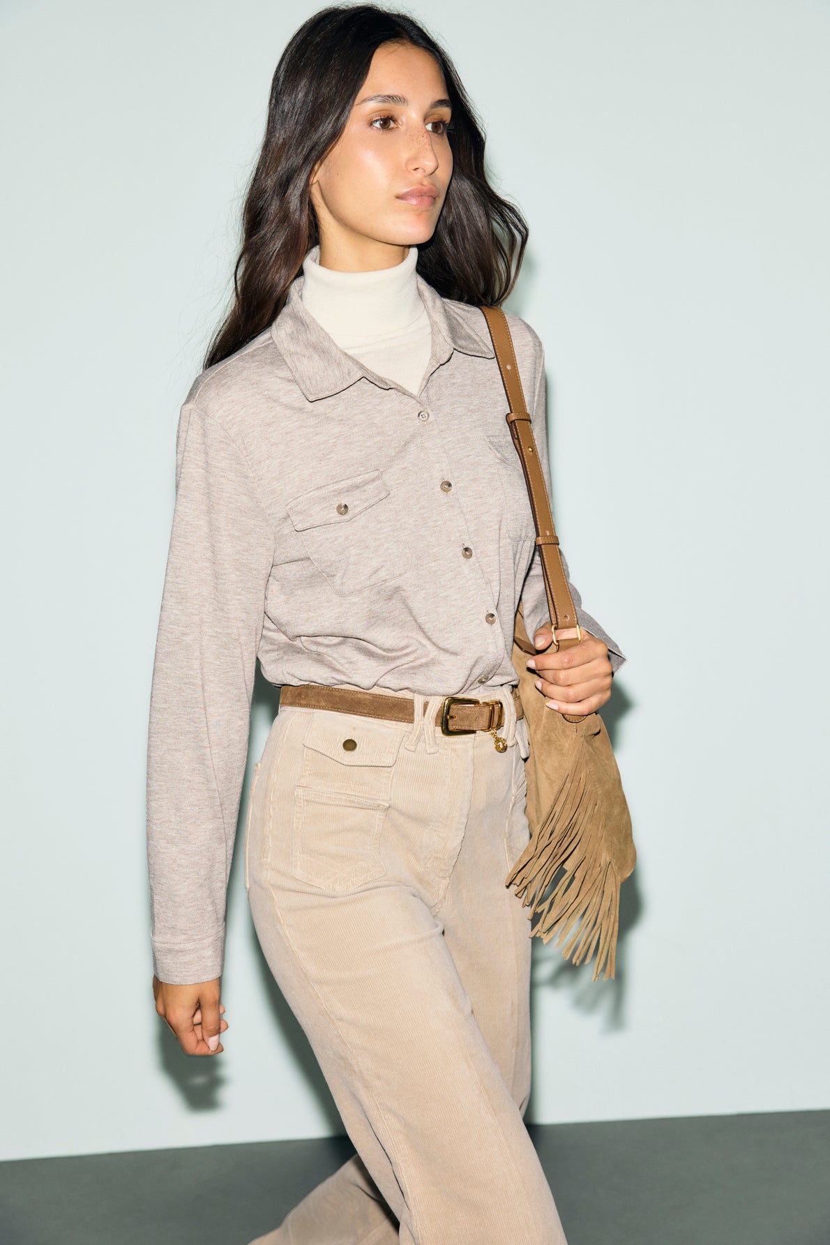 A woman with long dark hair wears a light turtleneck, button-up shirt with poches à la poitrine, beige high-waisted pants, and a brown fringed shoulder bag, walking against a pale blue background.