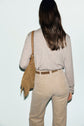 A person with long brown hair, seen from behind, wears a light beige tee-shirt beige with poches à la poitrine, matching pants, and carries a tan fringed shoulder bag.