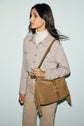 A woman with long dark hair wears a beige button-up shirt with poches à la poitrine, a light turtleneck, and tan pants. She carries a tan suede fringe shoulder bag and stands against a light blue background, looking to the side.