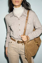 Woman wearing a beige button-up shirt with poches à la poitrine over a white turtleneck, light tan corduroy pants, and a brown belt, holding a tan suede fringe shoulder bag. Her face is partially out of the frame.