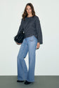 Woman stands against a plain background wearing a dark gray pull femme en maille côtelée avec col bateau, light blue wide-leg jeans, and holding a large black handbag. She has long brown hair and a relaxed expression.