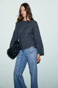 Woman wearing a dark gray pull femme à col bateau avec des détails de boutons et confectionné en maille côtelée, associé à un jean bleu clair, tient un sac à main noir. Elle se tient devant un fond uni et clair.