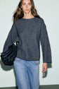 A woman with long brown hair wears a dark gray pull femme en maille côtelée à col bateau and light blue jeans, holding a black bag. She stands against a pale, neutral background.