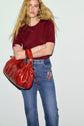 A woman with long blonde hair wears a red pull col rond en laine mérinos and blue jeans with patches, holding a large red bag. She stands against a plain light background with her arms folded.
