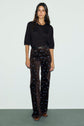 Woman standing against a plain background wearing a black laine mérinos pull col rond and brown high-waisted pants with blue floral patterns, accessorized with a black belt and brown shoes.