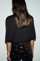 A person with long brown hair, wearing a pull col rond noir à manches courtes and dark pants with small blue flower patterns, is standing facing away from the camera against a light background.