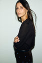 A woman with long dark hair stands sideways, arms crossed, wearing a pull col rond à manches courtes in soft laine mérinos and dark pants with small blue designs, against a plain light-colored background.