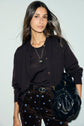 A woman with long brown hair wears a black cardigan, black shirt, and brown pants with small blue flower prints, carrying a black handbag and wearing a necklace, standing against a plain light background. Her look is elegantly finished with a classic gilet boutonné for added style.