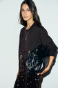 A woman with long dark hair wears a gilet marron and patterned pants, holding a large black leather bag and looking at the camera against a light background.