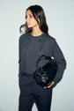 Woman with long dark hair wears a charcoal gilet laine mérinos cardigan and matching pants, holding a black leather handbag with gold hardware, looking to the side against a light gray background.