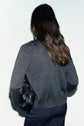 A person with long brown hair, wearing a gray gilet laine mérinos and dark pants, stands facing away, holding a black handbag under their arm against a plain light background.