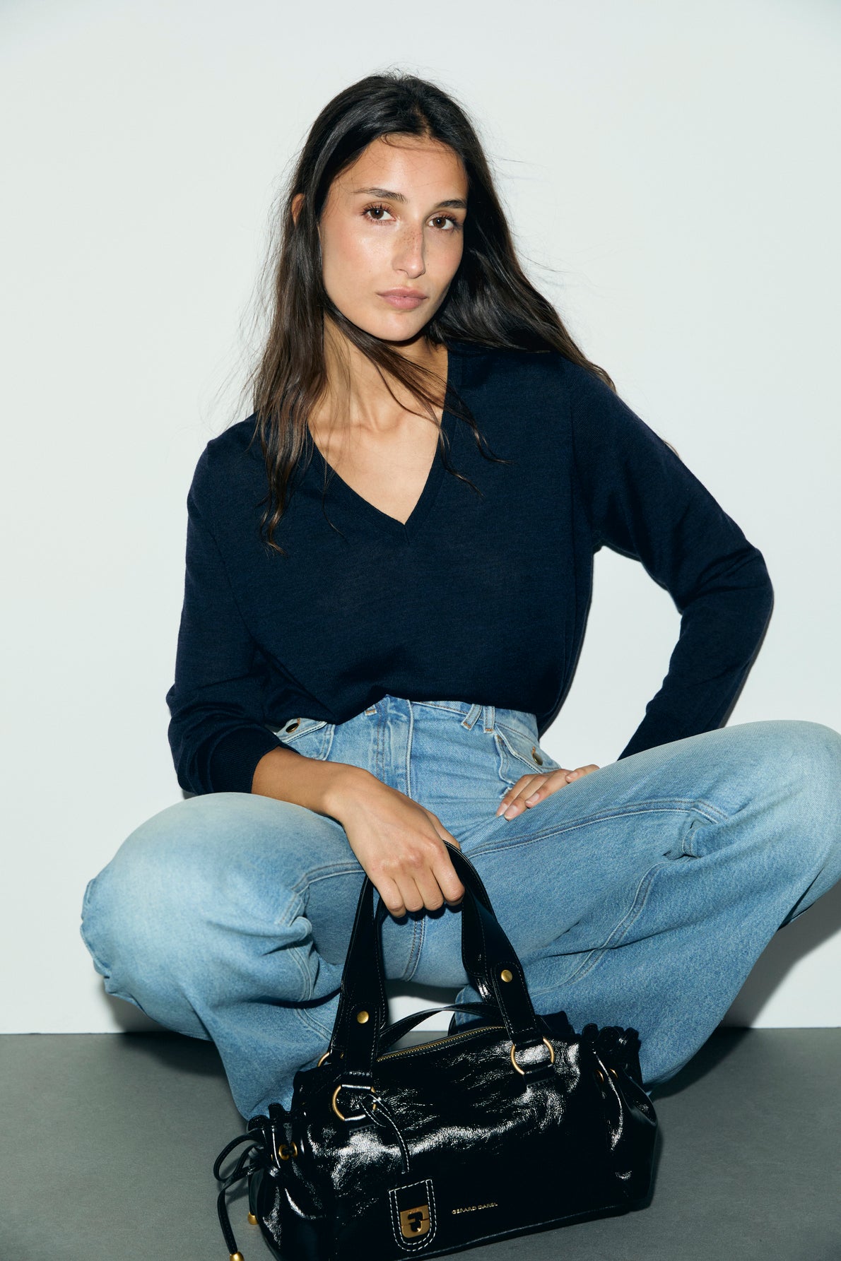 A woman with long dark hair sits cross-legged in light blue jeans and a navy sweater reminiscent of a pull bleu marine homme, holding a black patent leather handbag against a plain white background.