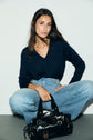 A woman with long dark hair sits cross-legged in light blue jeans and a navy sweater reminiscent of a pull bleu marine homme, holding a black patent leather handbag against a plain white background.