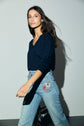 A woman with long brown hair wearing a navy long-sleeve top reminiscent of a pull bleu marine homme and light blue jeans with patches walks confidently, looking slightly to the side, against a plain light gray background.