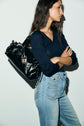 Woman with long brown hair wearing a pull col V laine mérinos navy top and high-waisted jeans, carrying a shiny black shoulder bag with gold hardware, standing against a plain white wall, arms crossed.