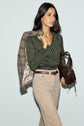 Woman with long dark hair wears a green pull kaki top, beige pants, and holds a brown bag. She drapes a plaid blazer over her shoulder. She has a neutral expression and stands against a light background.