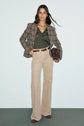 A woman stands against a light background wearing a plaid blazer, a pull col V laine in green, beige wide-leg pants, brown boots, and holding a brown handbag.