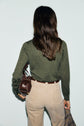 A person with long brown hair, seen from behind, wears a pull col V laine in dark green and beige pants, holding a brown leather bag and a plaid garment against a light background.