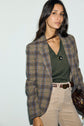 Woman wearing a plaid blazer, green top, beige pants, and a necklace—layered with a cozy pull col V laine—holding a brown bag, stands against a plain light background.