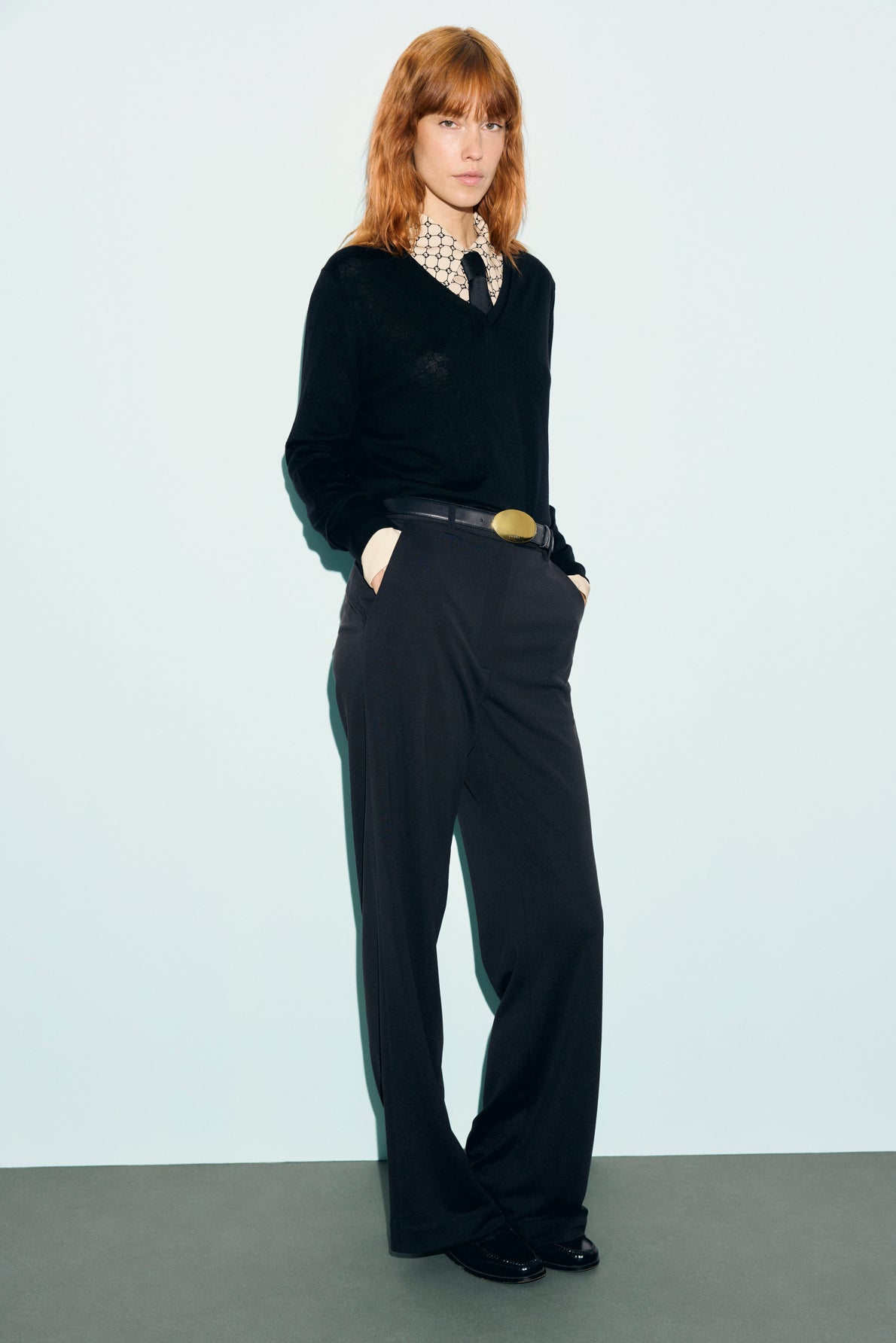 Person with red hair stands against a light background, wearing a pull en laine mérinos noir à col V, a patterned collared shirt, black wide-leg pants with a belt, and black shoes, with hands in pockets.