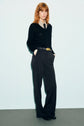 Person with red hair stands against a light background, wearing a pull en laine mérinos noir à col V, a patterned collared shirt, black wide-leg pants with a belt, and black shoes, with hands in pockets.