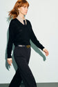 A woman with red hair walks confidently, wearing a pull en laine mérinos noir à col V superposé sur une chemise à motifs à col, un pantalon noir et une ceinture noire à boucle dorée, sur un fond clair et uni.